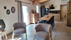 Uma sala de estar espaçosa com teto alto, vigas de madeira, mesa de jantar com cadeiras, área de cozinha e um espaço de estar com duas poltronas.