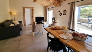 Uma sala de estar ampla, com teto de madeira, um sofá, uma televisão e uma mesa de jantar posta para dois.