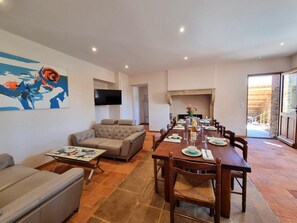 Ein Wohnzimmer mit einer Couch, einem Couchtisch und einem an der Wand befestigten Fernseher. Es gibt einen Essbereich mit einem langen Tisch, gedeckt für sechs Personen, umgeben von Stühlen.