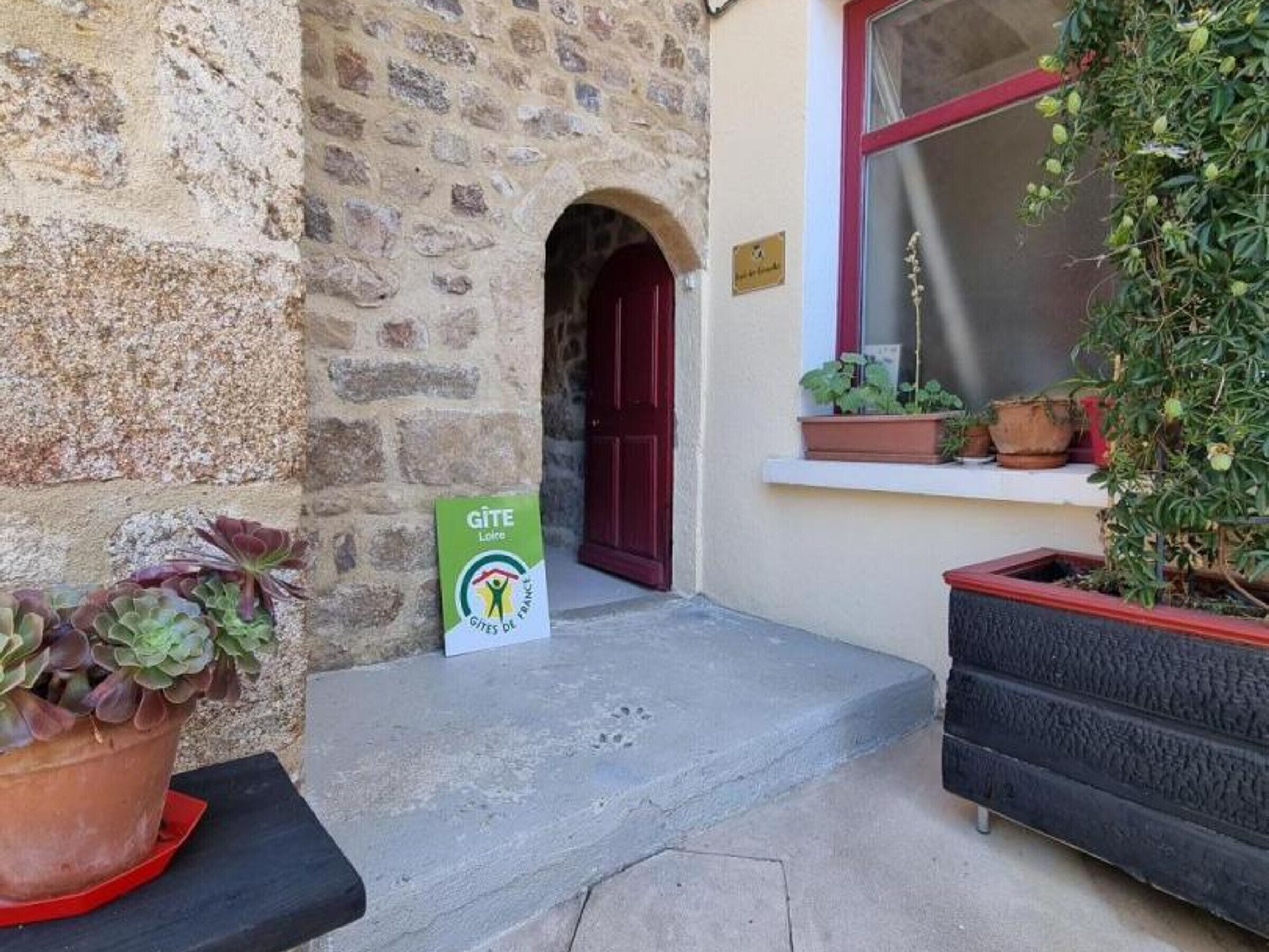 Un bâtiment en pierre avec une porte rouge, une fenêtre ornée de plantes en pot et une enseigne.