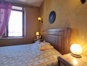 A bedroom with a bed, a nightstand, a wall clock, and a window with curtains. - Authentic house in the village center - 6 guests (ROZIER COTES D'AUREC)