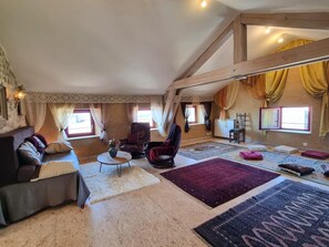 A spacious loft-style room with a high ceiling, exposed wooden beams, a sofa, a chair, a small table, and a variety of rugs. - Authentic house in the village center - 6 guests (ROZIER COTES D'AUREC)