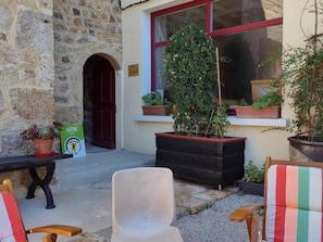 A stone building with a red door, a window with a plant, and outdoor seating. - Authentic house in the village center - 6 guests (ROZIER COTES D'AUREC)