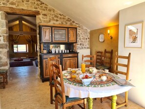 A dining area with a round table set for breakfast, wooden chairs, a stone wall, and a kitchen area with cabinets. - Authentic house in the village center - 6 guests (ROZIER COTES D'AUREC)