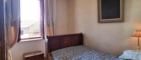 A bedroom with a wooden bed, a small bedside table, a window with curtains, and a wall-mounted picture frame.
