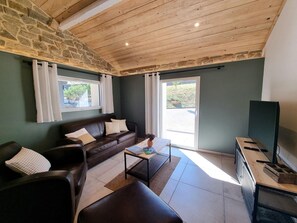 A living room with a stone wall, wooden ceiling, a brown leather sofa, a coffee table, and a television on a stand. - Peaceful air-conditioned lodge - 3 guests (Saint-Just-Saint-Rambert)