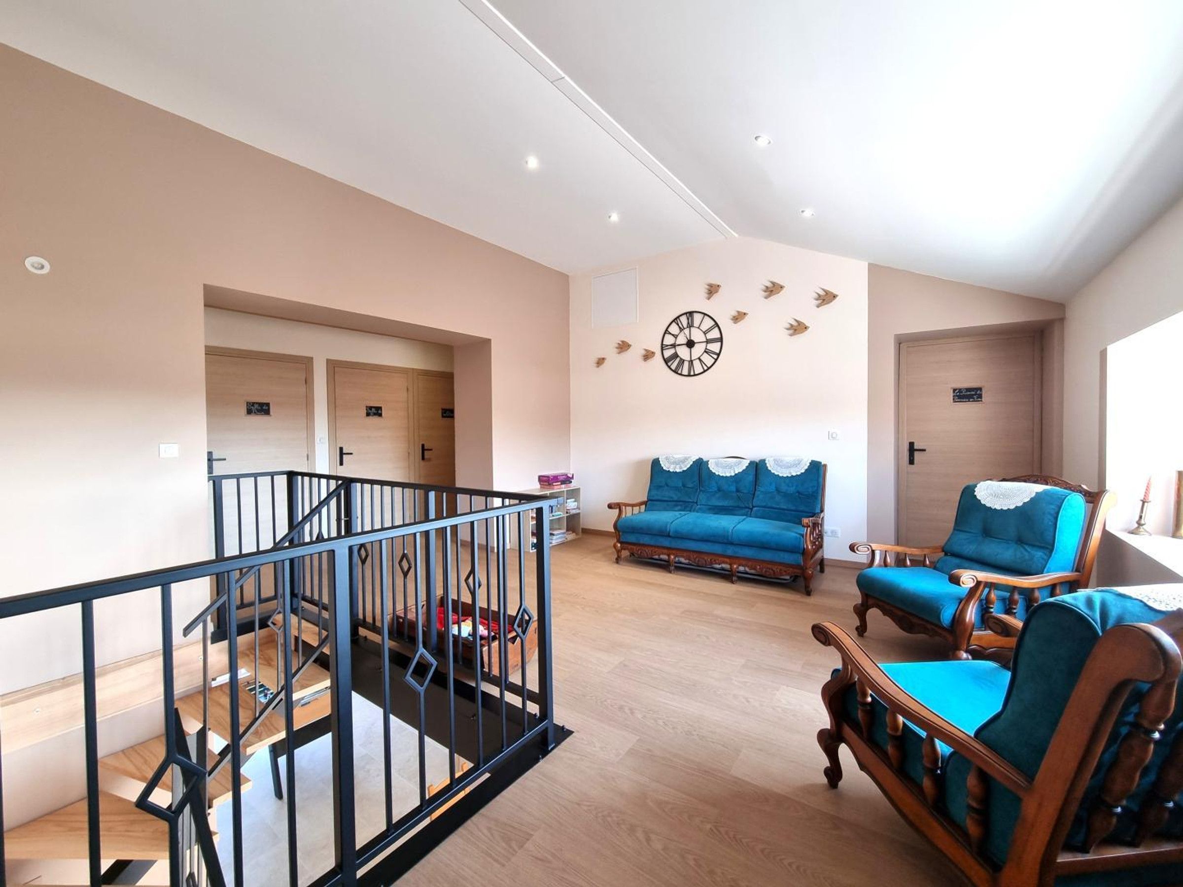 Een ruime woonkamer met een blauwe bank, houten stoelen, een trap met een zwarte leuning en een wandklok.