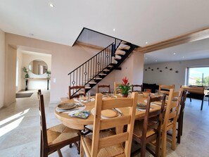 Un comedor con una mesa y sillas de madera, una escalera con barandilla negra, y un amplio ventanal que proporciona luz natural.