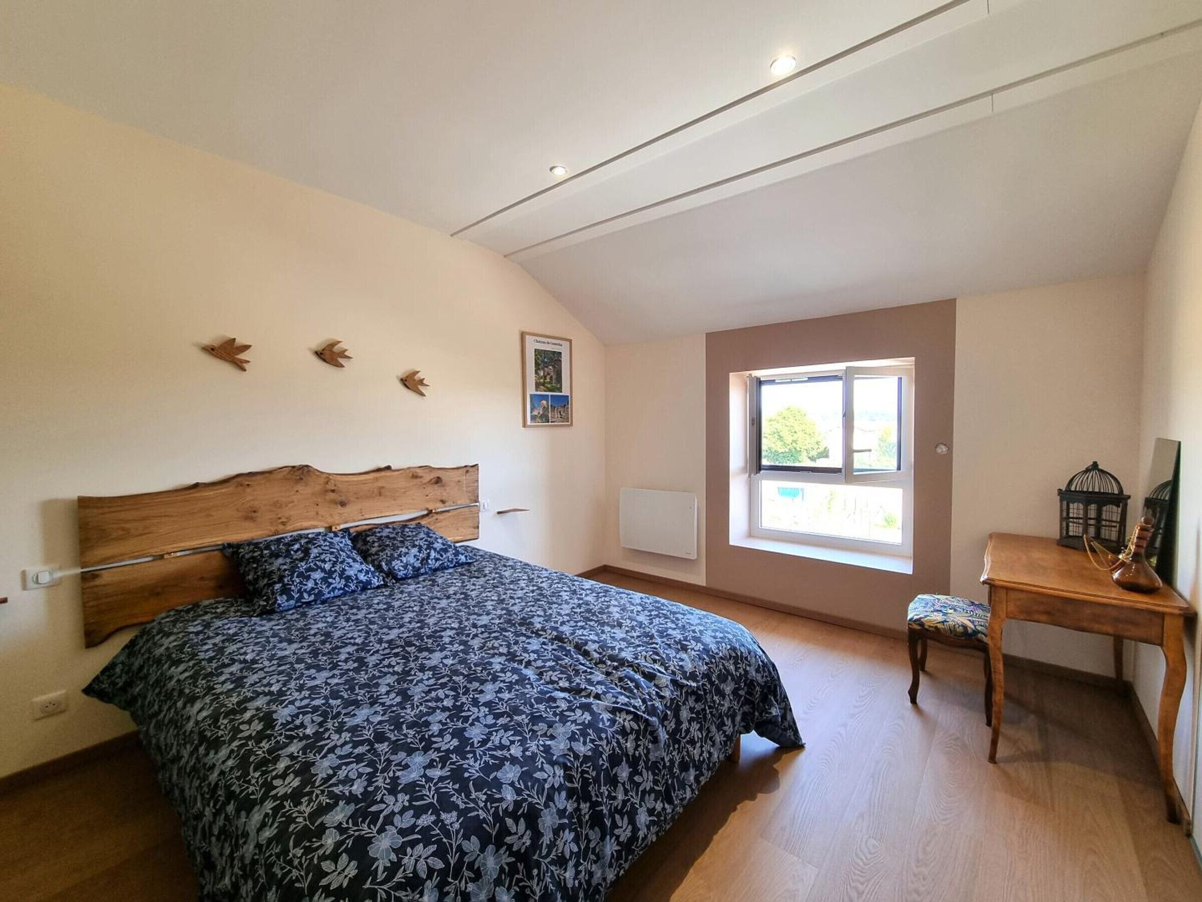 Une chambre avec une tête de lit en bois, un lit recouvert d’une couvre-lit à motifs floraux, une fenêtre donnant sur un beau paysage et une petite table avec une lampe.