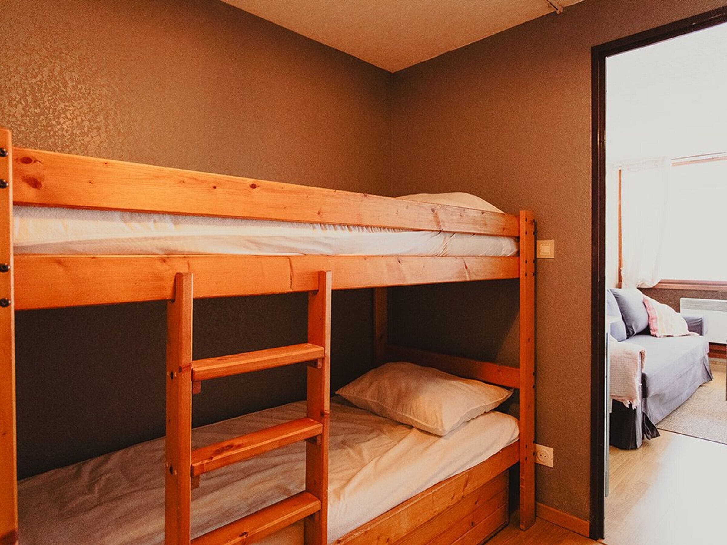 A bunk bed with a ladder, a single bed, and a small shelf unit in a room with a plain wall and a door leading to another room.