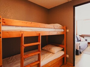 A bunk bed with a ladder, a single bed, and a small shelf unit in a room with a plain wall and a door leading to another room. - Apartment in the heart of the village - 4 guests (Saint-Lary-Soulan)