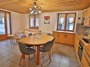 A cozy room with a wooden ceiling, a round dining table with chairs, a sofa, and a kitchen area with wooden cabinets and a window. - Apartment with Wi-Fi and parking - 6 guests (Les Gets)