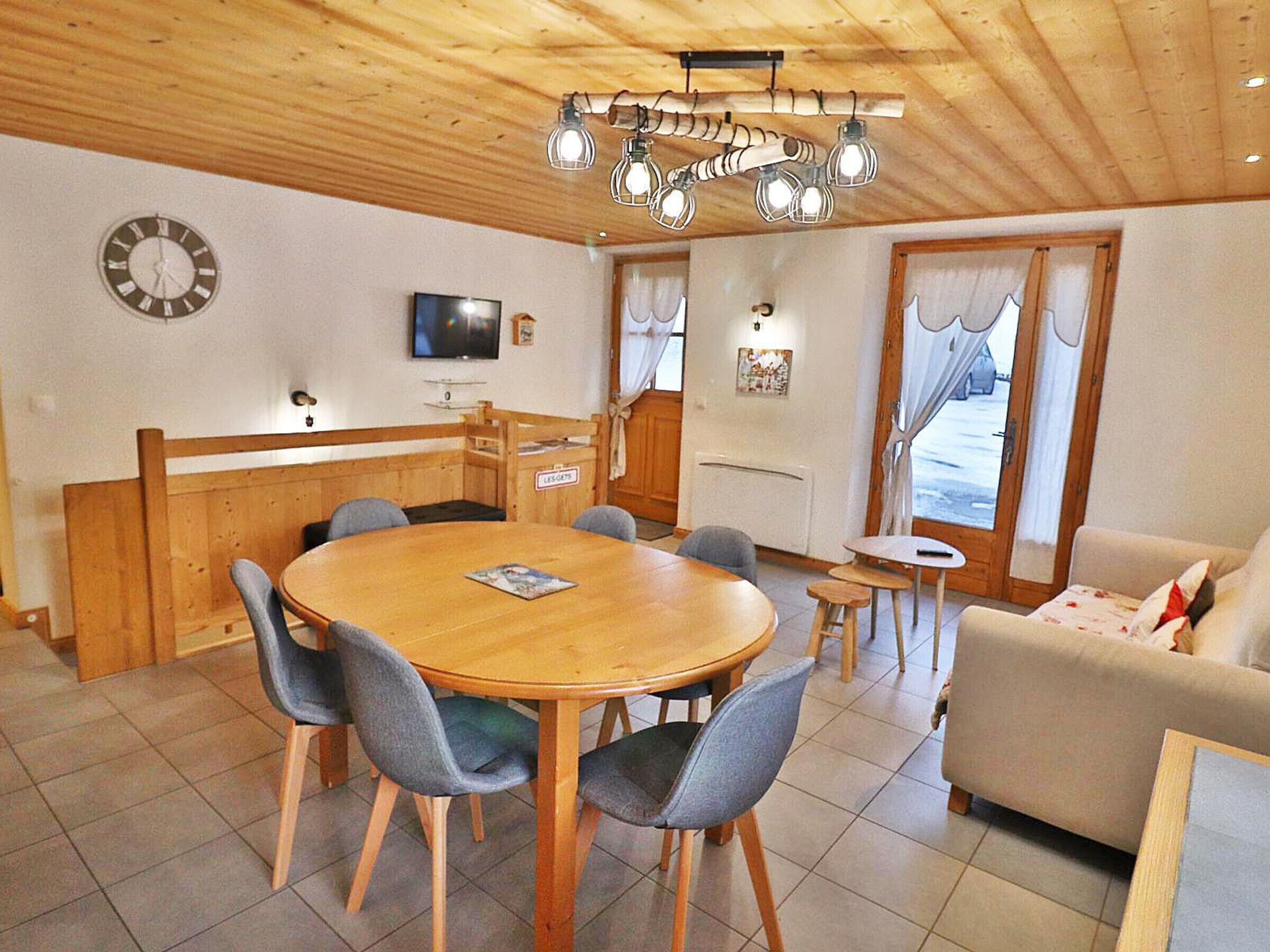A dining area with a wooden table and chairs, a television on the wall, and a sofa in the corner.