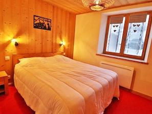 A bedroom with a wooden headboard, a bed with white bedding, two wall-mounted lights, a window with curtains, and a radiator. - Apartment with Wi-Fi and parking - 6 guests (Les Gets)