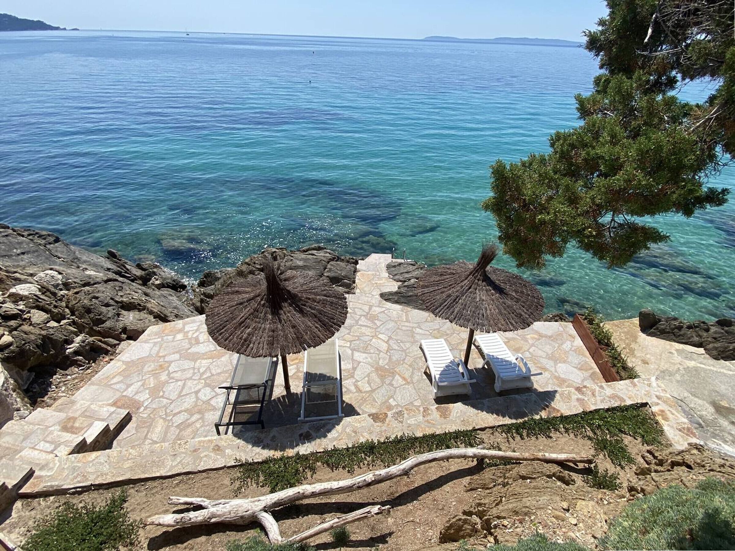A stone patio with two lounge chairs and thatched umbrellas overlooking a clear sea.