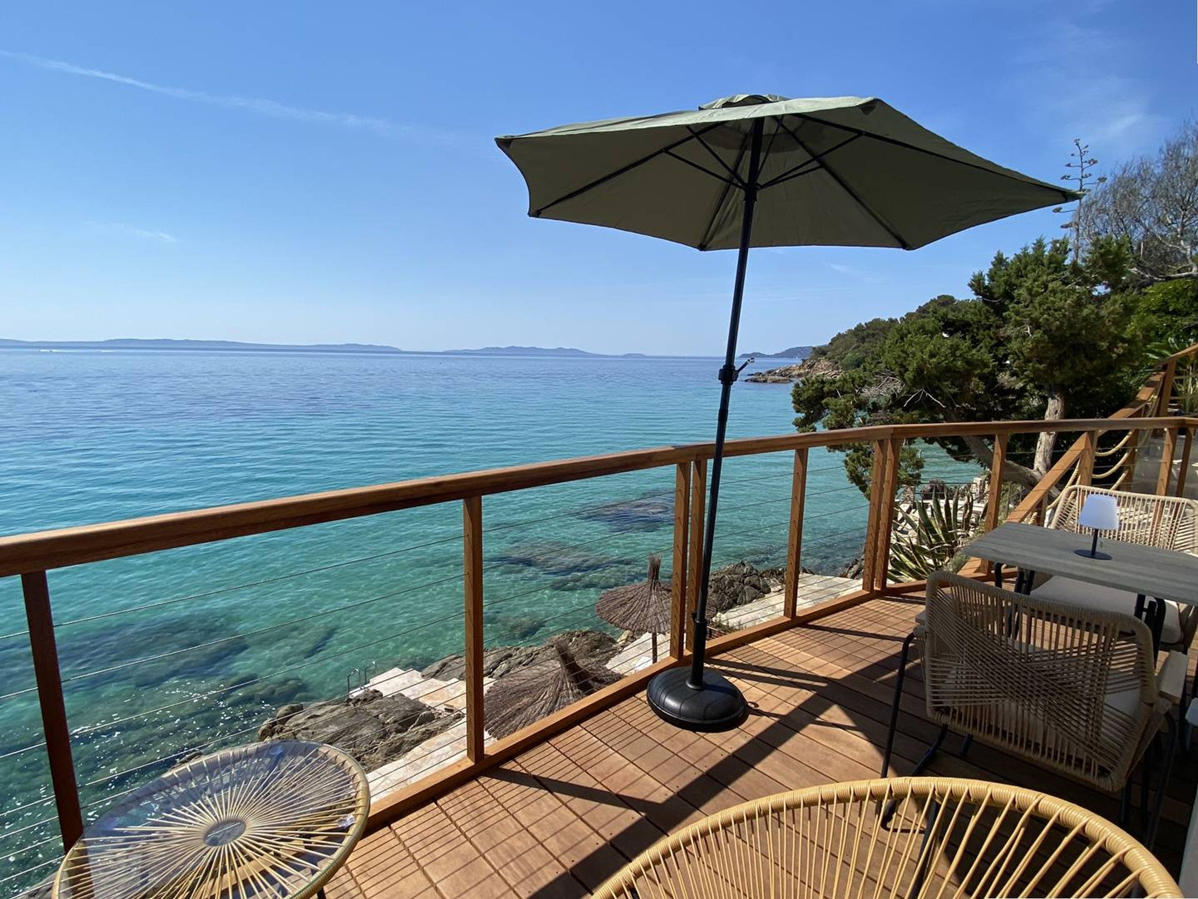 Una terraza de madera con vistas despejadas al mar, una silla de mimbre, una mesa y una sombrilla.