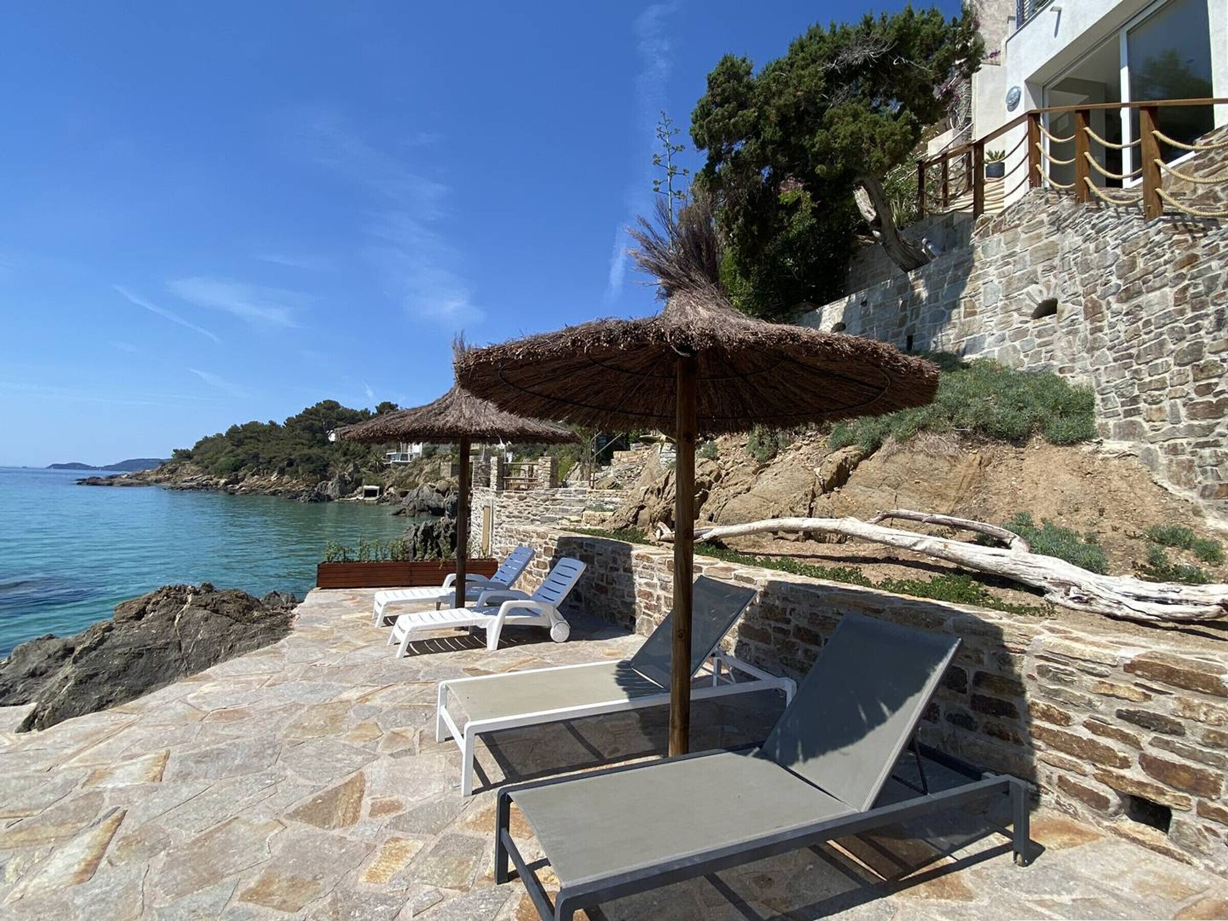 Un patio de piedra con sillones de descanso y un toldo de paja, con vista a una zona costera con un edificio de piedra y cielo despejado.