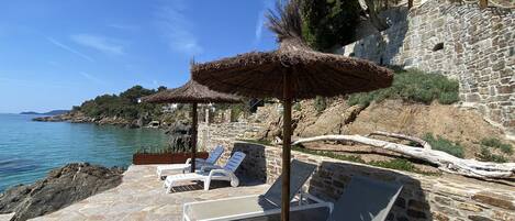 A stone patio with lounge chairs and a thatched umbrella, overlooking a coastal area with a stone building and a clear sky.