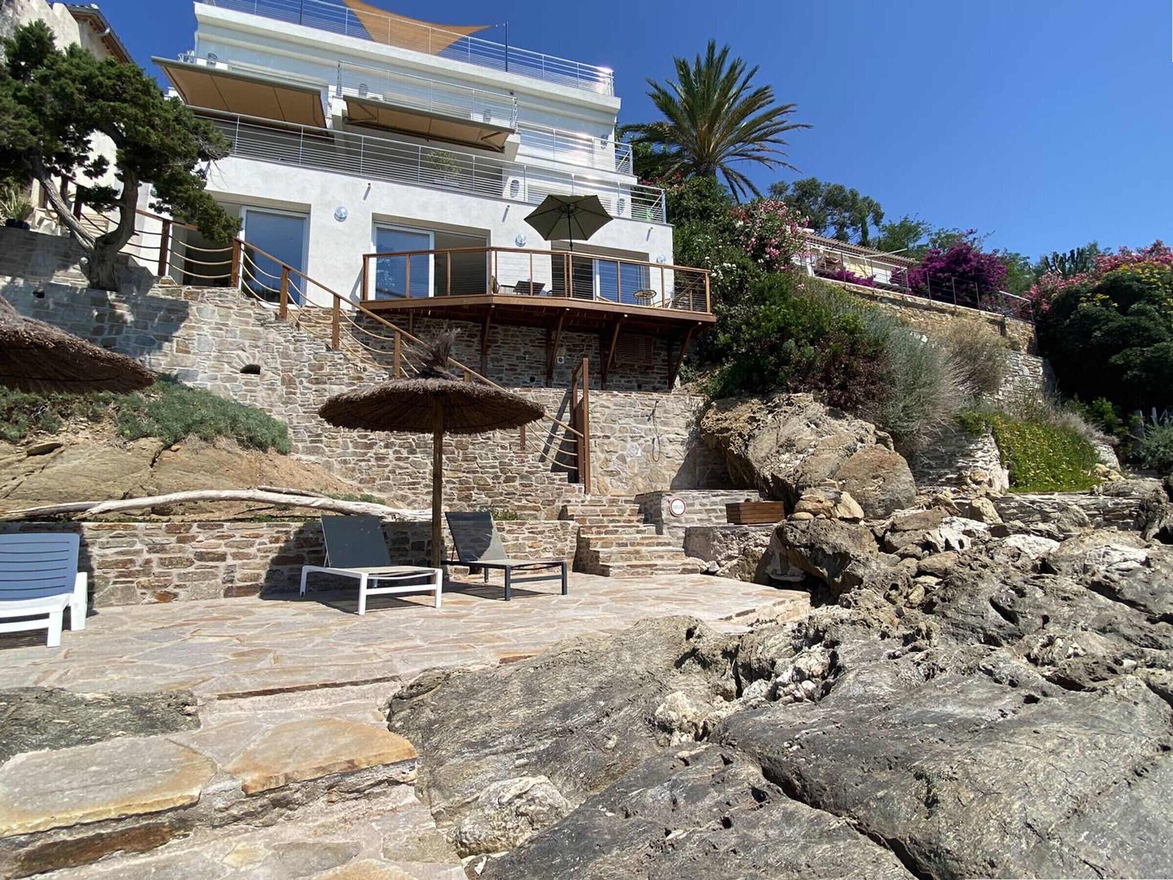 A stone building with a balcony, a thatched umbrella, and a seating area with chairs.