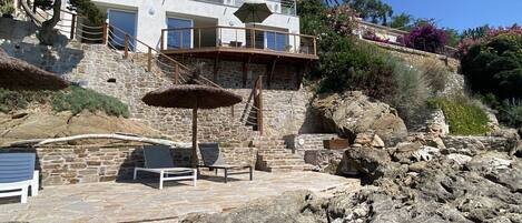 Edifici de pedra amb un balcó, una ombra de palla i una zona d'estar amb cadires.