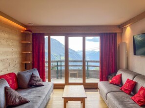 Une pièce au décor intérieur en bois, comprenant un ensemble de canapés, une table et une grande porte-fenêtre coulissante donnant sur un balcon avec vue sur la montagne.