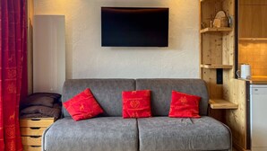 Ein gemütlicher Wohnbereich mit grauem Sofa, roten Kissen, einem Flachbildfernseher, Holzregalen und einer kleinen Küchenzeile.