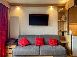 Ein gemütlicher Wohnbereich mit grauem Sofa, roten Kissen, einem Flachbildfernseher, Holzregalen und einer kleinen Küchenzeile.