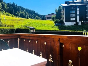 A wooden deck with heart-shaped cutouts, a white table, and a view of a green hillside with buildings in the background. - Comfortable apartment, ski-in access - 4 guests (Les Gets)