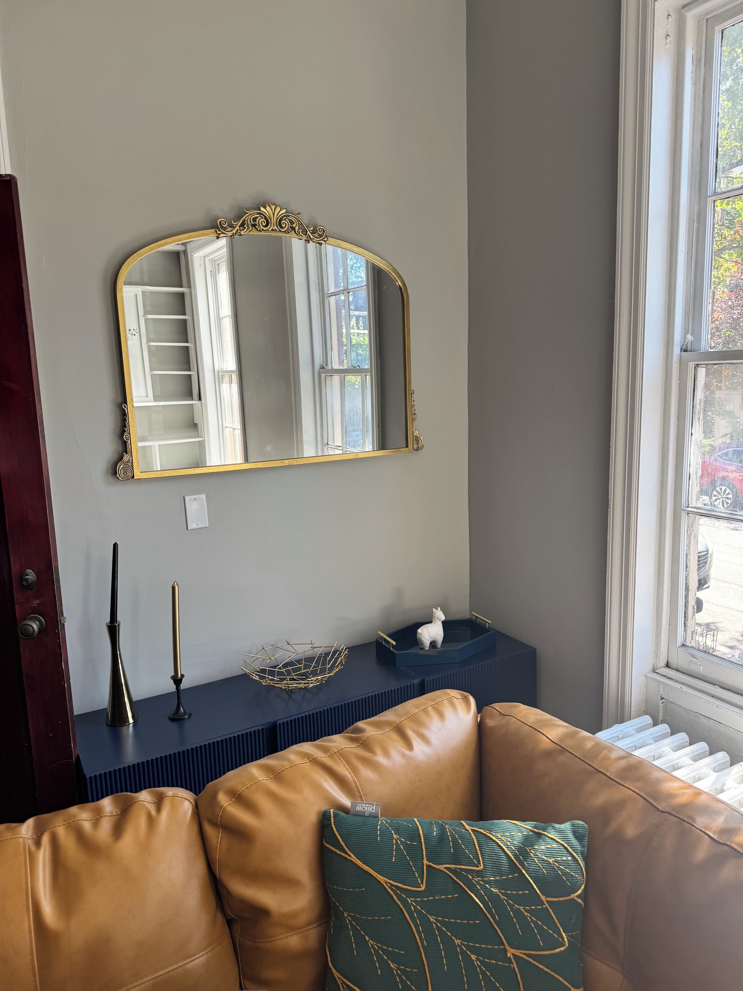 Une pièce dotée d’un grand miroir, d’un canapé en cuir marron, d’une petite table ornée d’objets décoratifs et d’une fenêtre donnant sur l’extérieur.