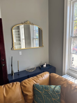 Ein Zimmer mit einem großen Spiegel, einem braunen Ledersofa, einem kleinen Tisch mit Dekoration und einem Fenster mit Blick ins Freie.