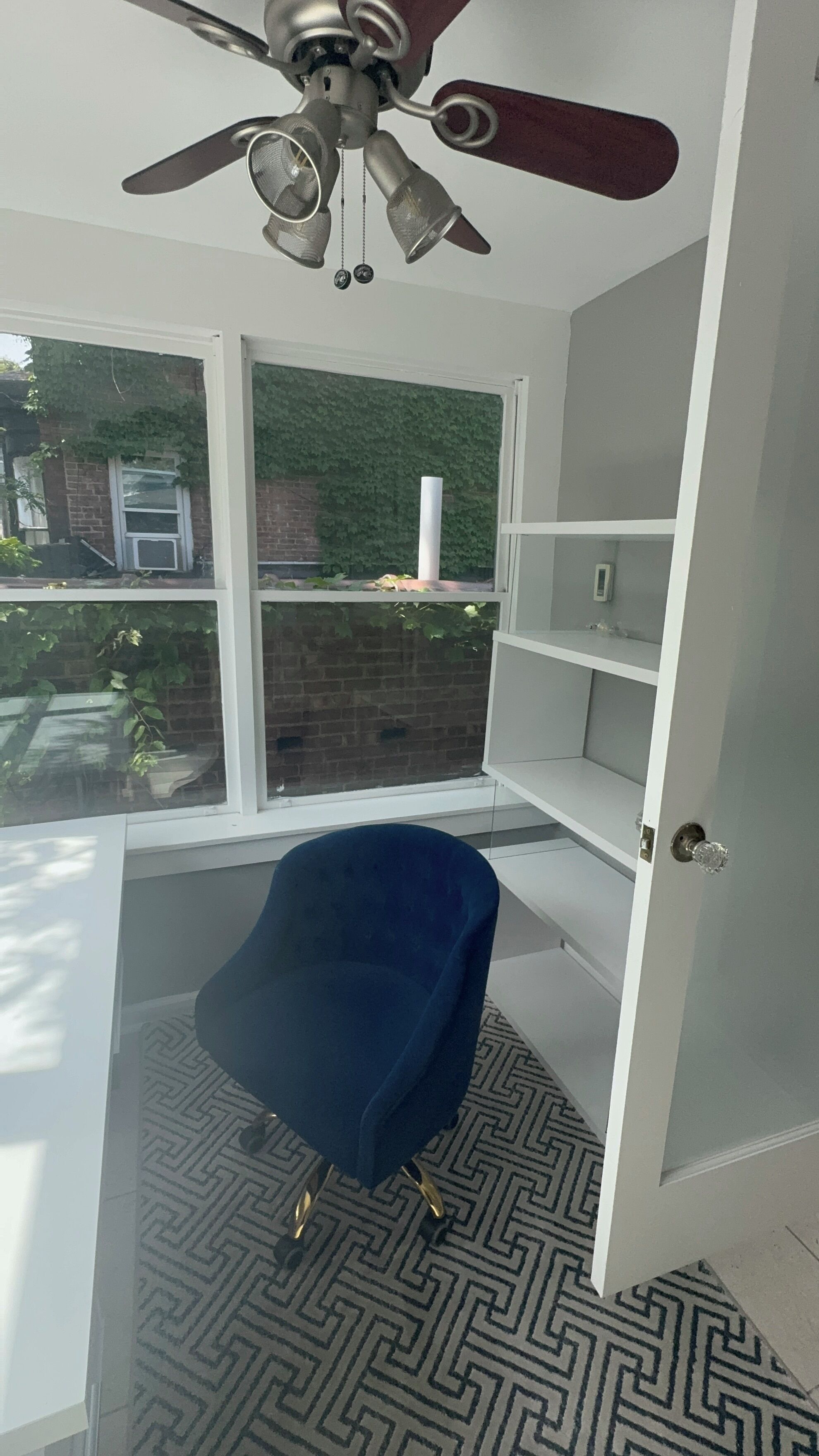 Une pièce dotée d’un ventilateur de plafond, d’une chaise bleue, d’un tapis à motifs et offrant une vue sur un mur de briques et sur la verdure à l’extérieur.