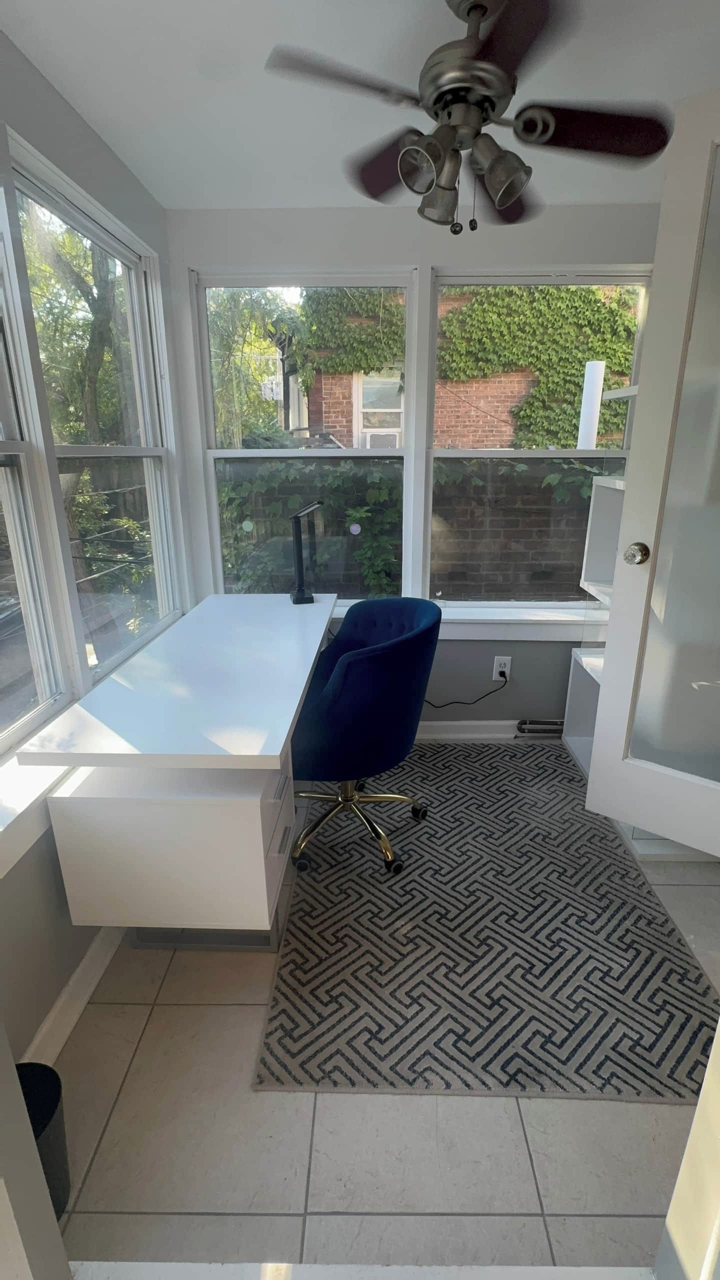 A room with a large window, a ceiling fan, a bathtub, a desk with a chair, and a patterned rug.