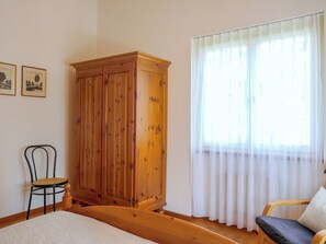 Ein Schlafzimmer mit einem Holzschrank, einem Bett, einem Sessel und einem Fenster mit transparenten Vorhängen.