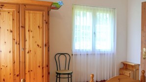 Quarto com guarda-roupa de madeira, uma cama, uma cadeira e uma janela com cortinas leves.