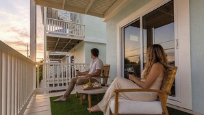 Ein Balkon mit zwei Personen, die auf Stühlen sitzen, einem Tisch mit einer Flasche und einer Speiseplatte, und einem Gebäude mit einem Balkon im Hintergrund.