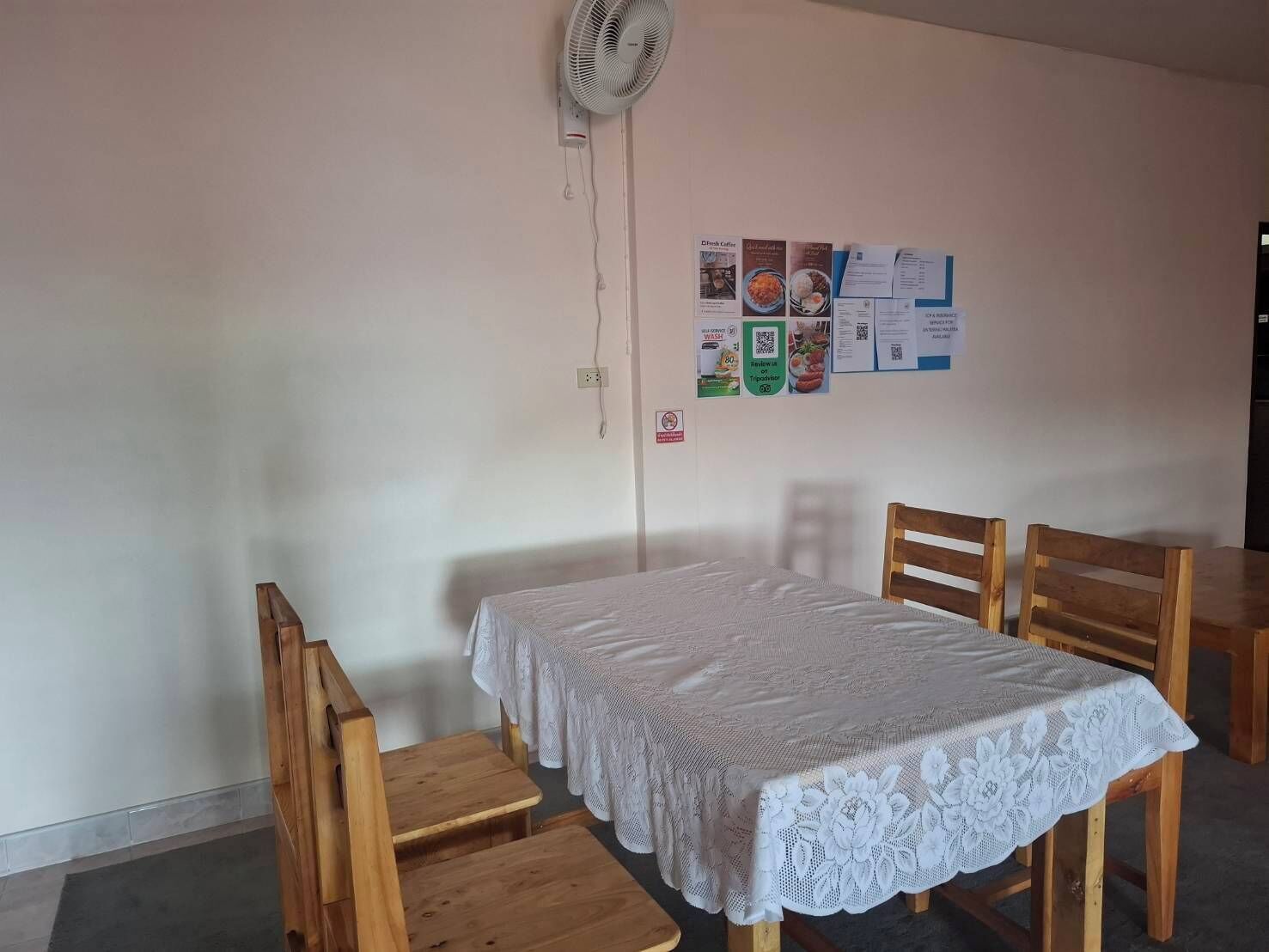 A room with a single table and two chairs, a white lace tablecloth, a wall-mounted fan, and a bulletin board with various papers.