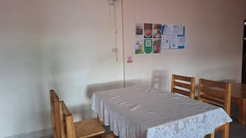 A room with a single table and two chairs, a white lace tablecloth, a wall-mounted fan, and a bulletin board with various papers.