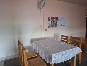 Una habitación con una mesa individual y dos sillas, un mantel de encaje blanco, un ventilador de pared y un tablón de anuncios con varios papeles.
