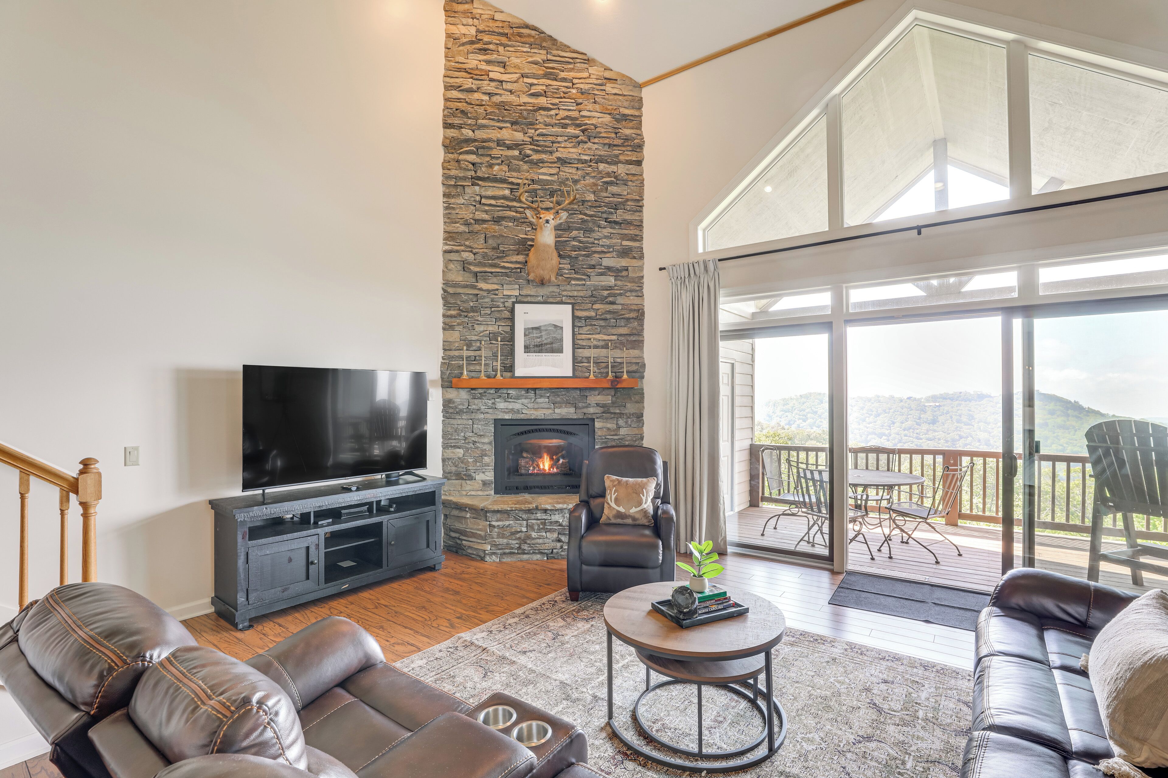 A spacious living room with a stone fireplace, a large flat-screen TV, leather sofas, and a balcony with outdoor furniture.