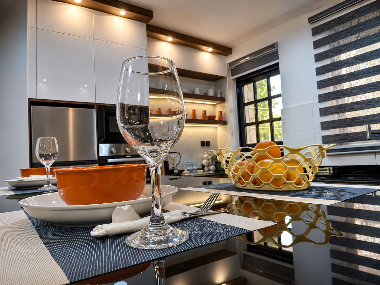 A modern kitchen with a dining table set for two, featuring a clear glass wine glass, a bowl, and a basket of oranges.