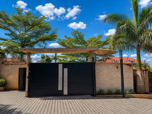 Una entrada protegida con un portón negro, una pérgola con toldo y un muro de ladrillo con palmeras.
