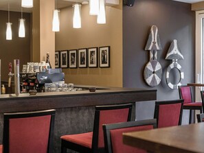 Un espace bar avec un comptoir, des verres et une machine à café. Des tableaux encadrés ornent le mur, ainsi qu’un élément décoratif mural.
