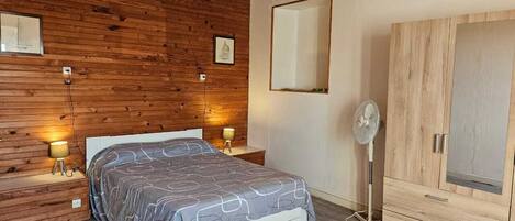 A bedroom with a wooden ceiling, a bed with a patterned bedspread, bedside lamps, a standing fan, and a wardrobe.