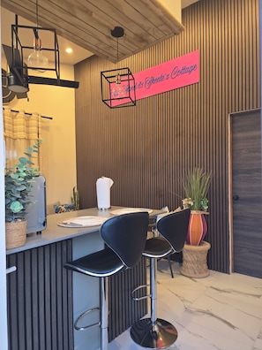 A modern bar area with a wooden slatted wall, a pink sign, black bar stools, and a potted plant. - Brilliant 1-bedroom apartment with AC in charming Oistins. Costline beaches near (Oistins)