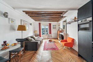 Ein modernes Wohnzimmer mit Fischgrätparkett, einer Couch, einem Esstisch mit Frühstück und einer Küchenzeile mit schwarzem Kühlschrank.