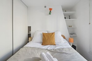 Ein modernes Schlafzimmer mit einem Bett, weißen Wänden, einer kleinen Ablage mit Dekoration und einer Pendelleuchte.