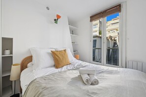 Ein ordentlich bezogenes Bett mit weißen Leinen und einem gefalteten Handtuch, ein braunes Kopfkissen und eine Nachttischlampe. Weiße Regale mit einigen Gegenständen darauf und ein Fenster mit Blick auf Gebäude.