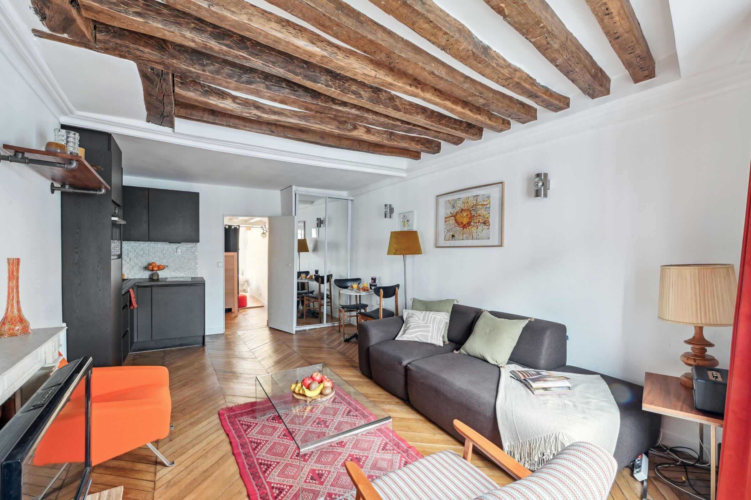 A modern living room with a wooden ceiling, a grey sofa, a glass coffee table, and a red patterned rug.
