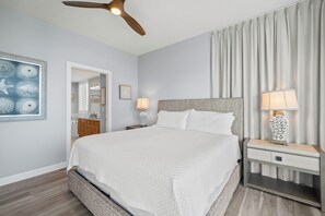 Ein Schlafzimmer mit einem Bett, Nachttischen, einem Deckenventilator und einem großen Fenster mit Vorhängen.