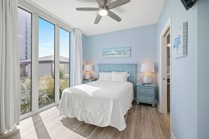 Ein Schlafzimmer mit einem Bett, zwei Nachttischen, einem Deckenventilator und Blick durch ein großes Fenster.